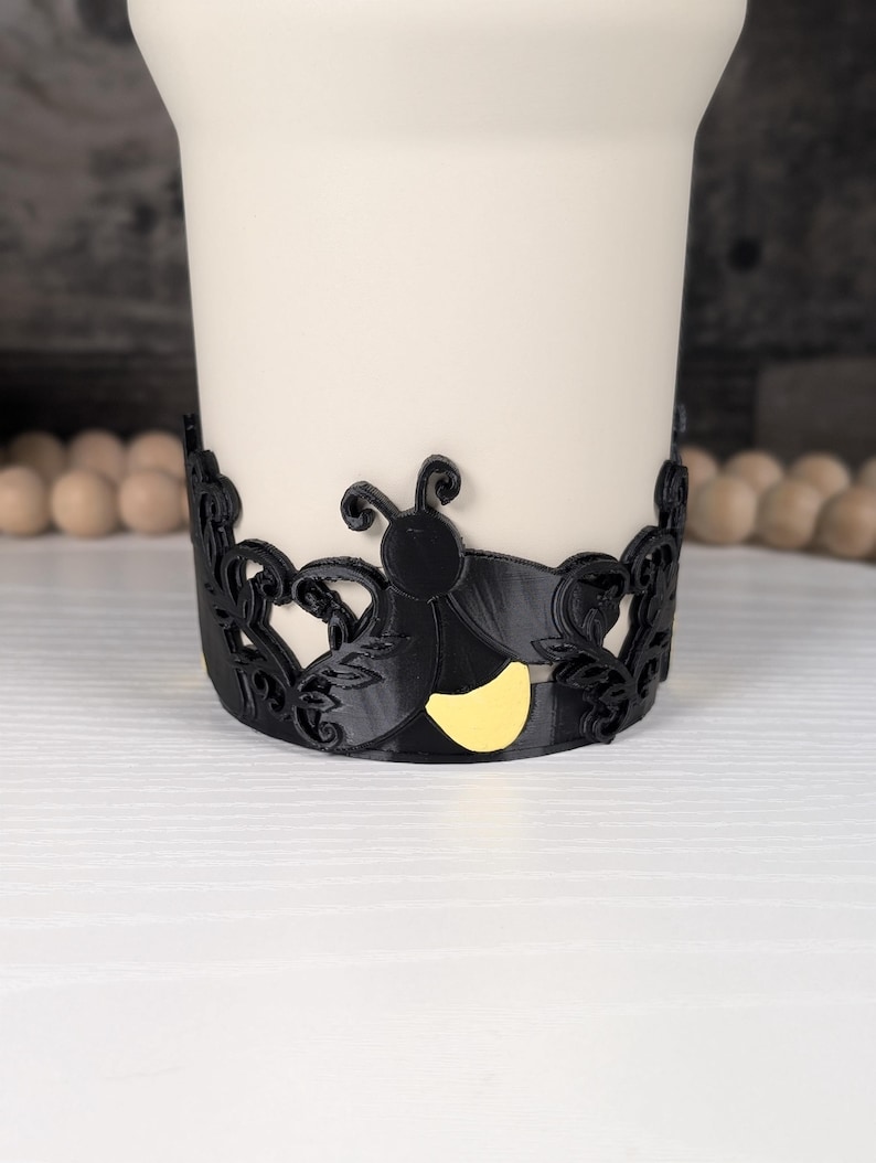 Puede incluir: Una banda decorativa negra que presenta la silueta de una luci&eacute;rnaga con un abdomen amarillo brillante, rodeada de intrincados patrones de hojas en filigrana. La banda est&aacute; colocada alrededor de un objeto cil&iacute;ndrico blanco liso, creando contraste.