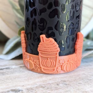 May include: A black tumbler with a leopard print design, paired with a peach-colored cup holder. The holder features a pumpkin spice latte design and pumpkin carvings, perfect for fall.