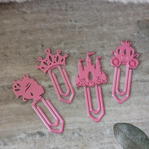 May include: Four pink metal paper clips with glitter. The clips feature princess-themed designs: a crown, a castle, a carriage, and a princess silhouette. The clips are arranged on a gray surface with decorative elements.