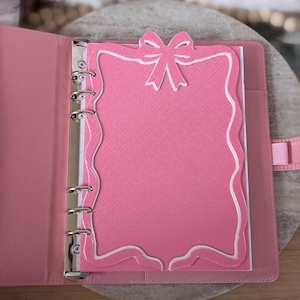 May include: Pink binder with a decorative pink and white paper insert. The insert has a bow at the top and a scalloped edge. The binder is open, revealing the metal rings and the inside cover.