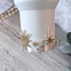May include: White tumbler with a gold glitter star design around the base. The stars vary in size and are attached to a white ribbon. The tumbler has a handle and a smooth, matte finish.