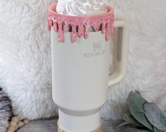 May include: A white Stanley travel mug with a pink, dripped-icing rim and a whipped cream-shaped lid. The mug has a handle and a beige base. A cherry and bow adorn the top. The Stanley logo is visible on the front.