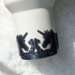 May include: A white tumbler with a black decorative band featuring two unicorn silhouettes. The band has an ornate, swirling design. The tumbler is set against a white, fluffy background.