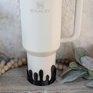 Puede incluir: Taza de viaje Stanley blanca con un logotipo de oso negro y la palabra "STANLEY" impresa en el lateral. La taza est&aacute; sobre un posavasos negro de estilo goteo.