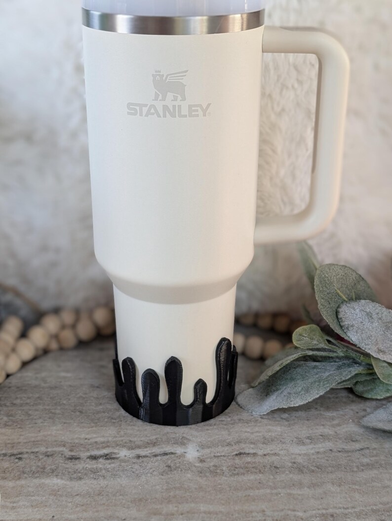 Puede incluir: Taza de viaje Stanley blanca con un logotipo de oso negro y la palabra "STANLEY" impresa en el lateral. La taza est&aacute; sobre un posavasos negro de estilo goteo.