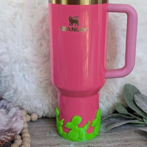 May include: A pink Stanley tumbler with a handle and a clear lid. The tumbler has a gold band and the Stanley logo. A green balloon dog design decorates the bottom. A pink straw is inserted into the lid.