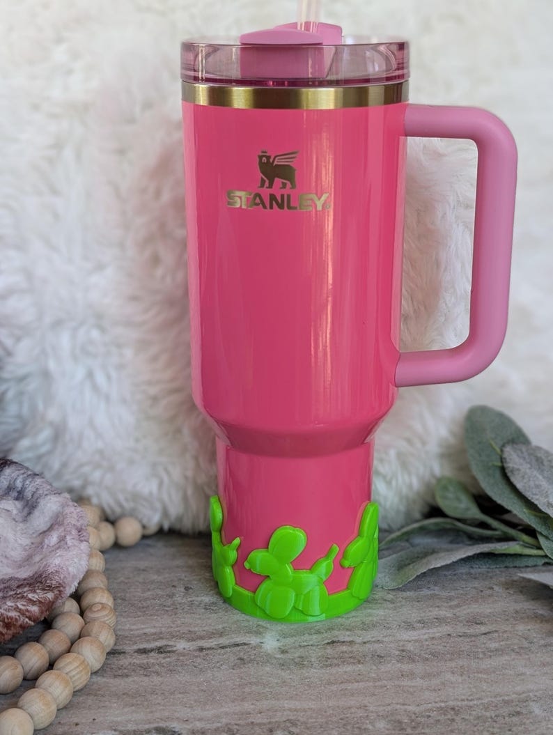 May include: A pink Stanley tumbler with a handle and a clear lid. The tumbler has a gold band and the Stanley logo. A green balloon dog design decorates the bottom. A pink straw is inserted into the lid.