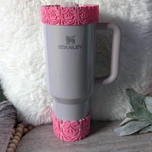 Puede incluir: Un vaso Stanley gris con bandas decorativas rosas en la parte superior e inferior, con un diseño ornamentado. El vaso tiene un asa y una pajita transparente. El logotipo de Stanley es visible en la parte delantera. Ideal para bebidas frías.