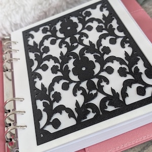 May include: A black, laser-cut floral design overlaying a white page within a pink binder. The intricate pattern features flowers and leaves. The binder has silver rings and a pink cover. The background is blurred, with a soft, white texture.
