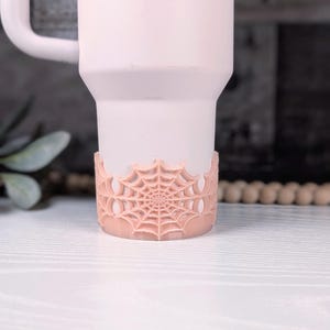May include: A light pink tumbler featuring a raised, textured spider web design wrapped around the base. The cup has a smooth matte finish and includes a matching light pink handle extending from the upper side of the cylindrical body.