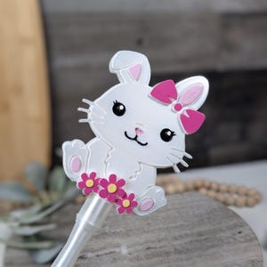 May include: A white bunny pen topper with a pink bow and pink flower accents. The bunny has black eyes and whiskers. The pen is white and has a ridged grip.