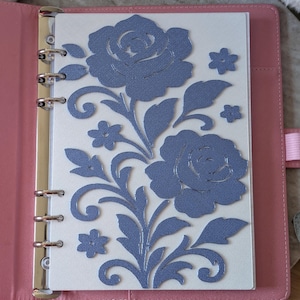 May include: A pink leather binder with a floral design. The design features two large blue roses and several smaller flowers and leaves. The binder is open, revealing the floral design on a white page.