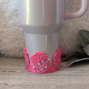 May include: A white tumbler with a pink glitter finish and a pink handle. The tumbler has a pink and silver glitter base with a checkered pattern.