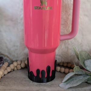 Puede incluir: Una taza de viaje Stanley rosa con un dise&ntilde;o de goteo negro alrededor de la parte inferior. La taza tiene una tapa dorada y un asa rosa.