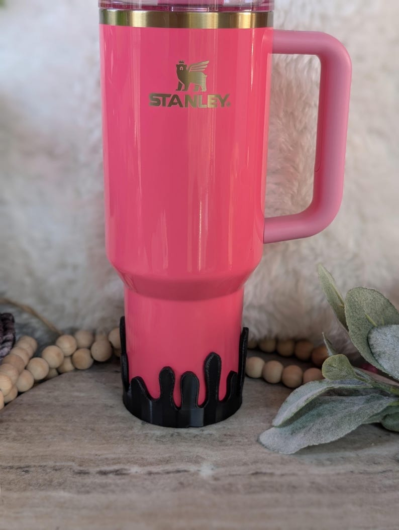 Puede incluir: Una taza de viaje Stanley rosa con un dise&ntilde;o de goteo negro alrededor de la parte inferior. La taza tiene una tapa dorada y un asa rosa.