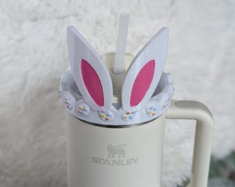 May include: White travel mug with a bunny ear lid and a pastel colored Easter egg design around the rim. The mug has a handle and the word "STANLEY" is printed on the side.