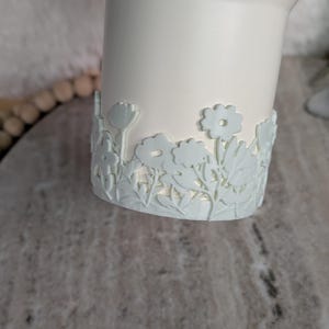 May include: A white ceramic vase with a light blue floral design around the base. The flowers and leaves are intricately detailed, creating a delicate and elegant aesthetic. The vase has a smooth, matte finish.