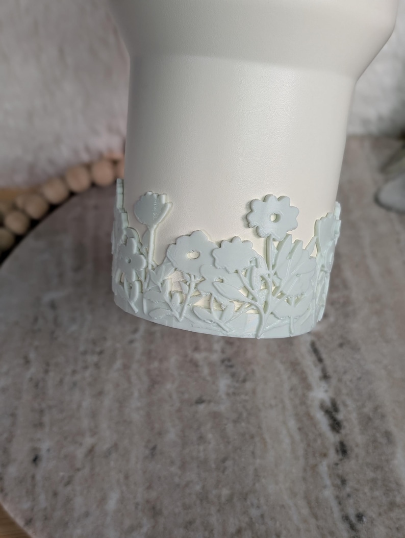 May include: A white ceramic vase with a light blue floral design around the base. The flowers and leaves are intricately detailed, creating a delicate and elegant aesthetic. The vase has a smooth, matte finish.