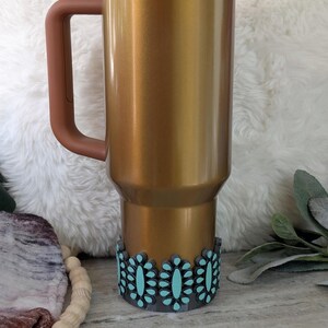 May include: A gold-colored insulated tumbler with a brown handle, lid, and decorative turquoise floral design at the base. The tumbler has a straw and is designed to keep beverages cold or hot. The tumbler is approximately 10 inches tall.