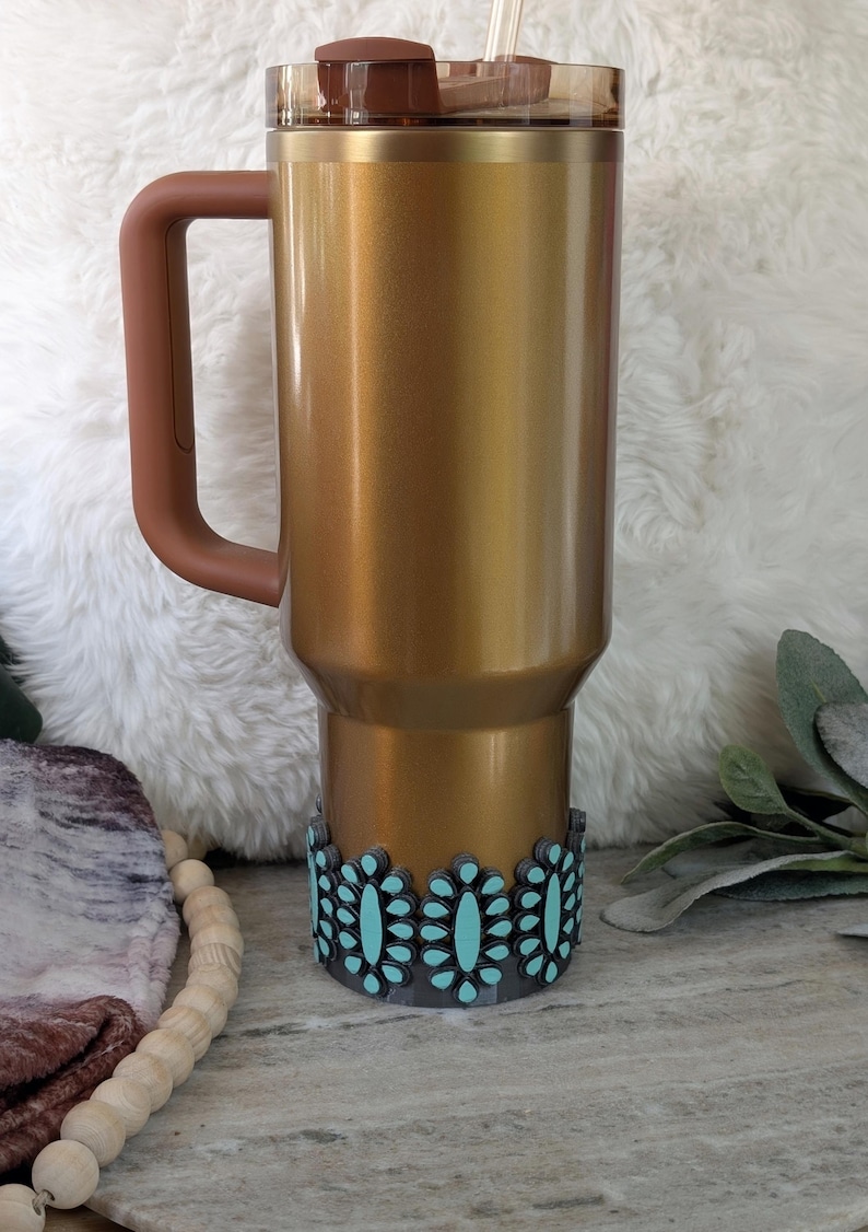 May include: A gold-colored insulated tumbler with a brown handle, lid, and decorative turquoise floral design at the base. The tumbler has a straw and is designed to keep beverages cold or hot. The tumbler is approximately 10 inches tall.