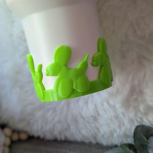 May include: A white tumbler with a bright green balloon animal design around the base. The balloon animals are in the shape of a dog. The tumbler sits on a white, fluffy surface.
