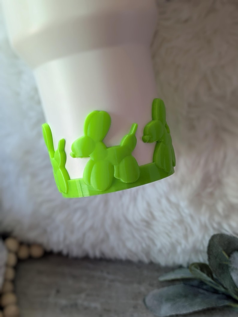 May include: A white tumbler with a bright green balloon animal design around the base. The balloon animals are in the shape of a dog. The tumbler sits on a white, fluffy surface.