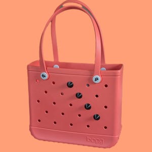 May include: Coral-colored tote bag with two handles and a rectangular shape. The bag features decorative black and silver accents and small circular perforations. The brand name "bogg" is printed on the bottom of the bag.