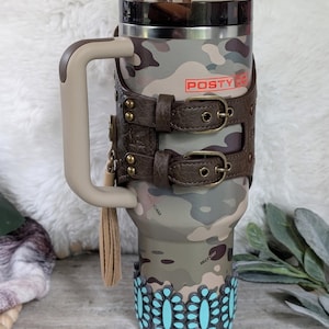 May include: A camouflage-patterned tumbler with a brown handle and a leather strap design. The tumbler features the text "POSTY CO" in red and is adorned with turquoise floral accents at the base. A brown straw is inserted into the lid.