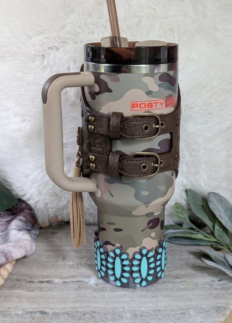 May include: A camouflage-patterned tumbler with a brown handle and a leather strap design. The tumbler features the text "POSTY CO" in red and is adorned with turquoise floral accents at the base. A brown straw is inserted into the lid.
