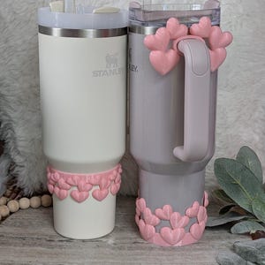 May include: Two insulated tumblers with straws. One is white, the other is gray. Both have pink heart-shaped decorations around the base and on the handle of the gray tumbler. The white tumbler has the "Stanley" logo.