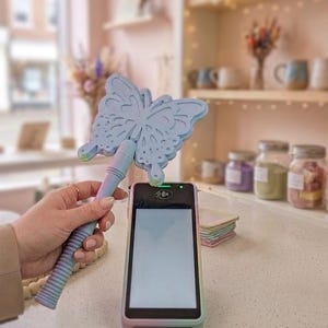 May include: A pastel-colored butterfly-shaped card reader accessory is held above a point-of-sale device. The accessory has a textured handle and a detailed butterfly design. The card reader has a rainbow-colored border.