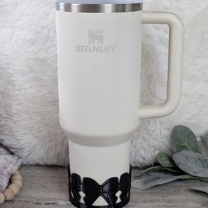 May include: A white Stanley travel mug with a black bow-shaped handle wrap. The mug has a silver lid and a stainless steel band around the middle. The Stanley logo is printed on the mug.