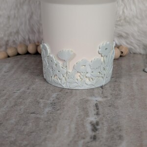 May include: A white ceramic vase with a floral design around the base. The flowers and leaves are in a raised relief pattern. The vase has a smooth, matte finish and a slightly flared top.