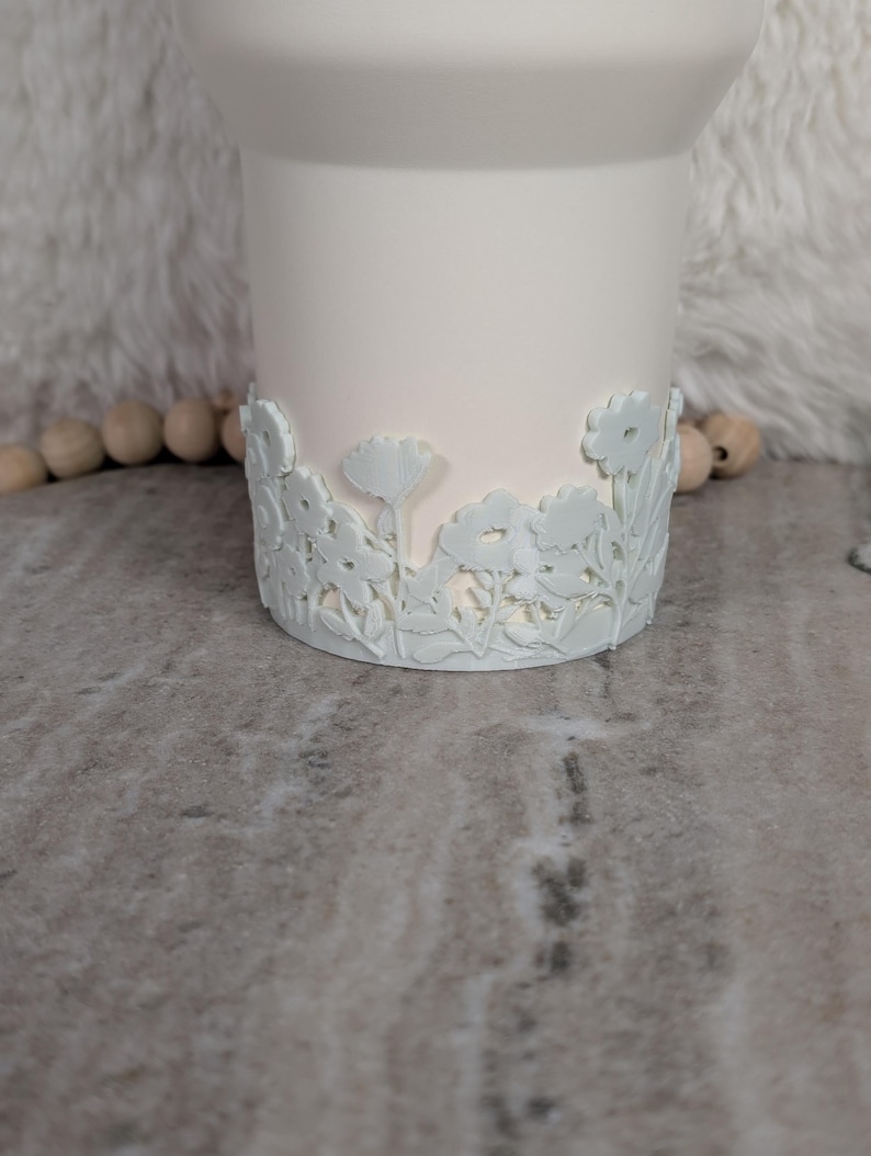 May include: A white ceramic vase with a floral design around the base. The flowers and leaves are in a raised relief pattern. The vase has a smooth, matte finish and a slightly flared top.