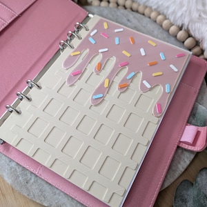 May include: A pink planner with a waffle-patterned interior and a clear cover featuring a pink, dripping design with colorful sprinkles. The planner is open, revealing the interior pages and a ring binder.