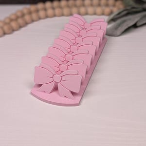 May include: A row of pink bow-shaped decorations. Each bow is detailed with multiple loops and a central knot. The bows are attached to a rectangular base with a curved edge. The color is a soft, pastel pink.