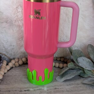 May include: A pink Stanley tumbler with a matching handle and lid. The tumbler has a gold rim and the Stanley logo. A green, drippy-style base is attached to the bottom. The tumbler is designed to hold a large amount of liquid.
