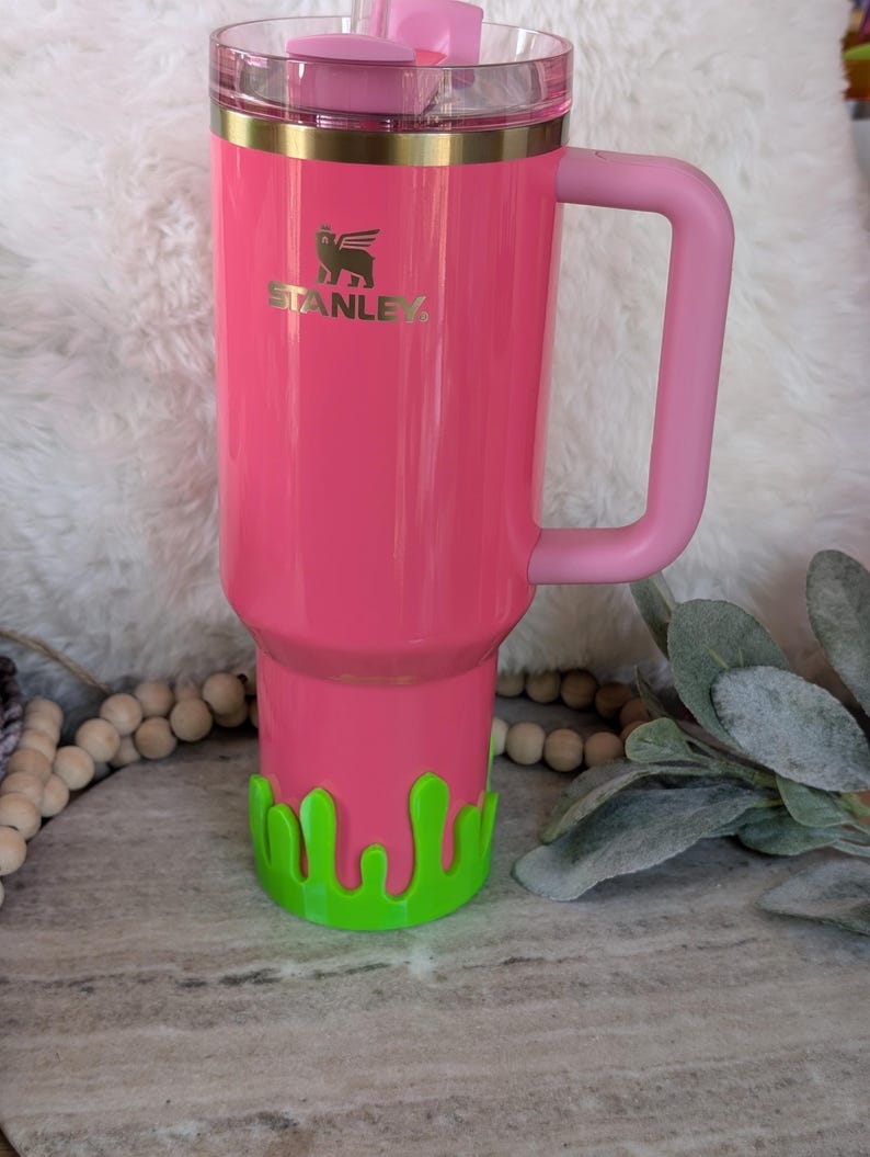 May include: A pink Stanley tumbler with a matching handle and lid. The tumbler has a gold rim and the Stanley logo. A green, drippy-style base is attached to the bottom. The tumbler is designed to hold a large amount of liquid.