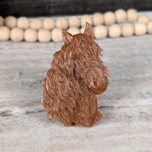 May include: A brown horse head sculpture with detailed mane and facial features. The sculpture is made of a textured material and is set against a neutral background. The horse head is a decorative item.