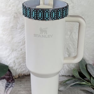 May include: A white Stanley tumbler with a handle and a decorative band featuring turquoise-colored stones. The tumbler has a straw and the Stanley logo. The overall design is clean and modern, suitable for beverages.