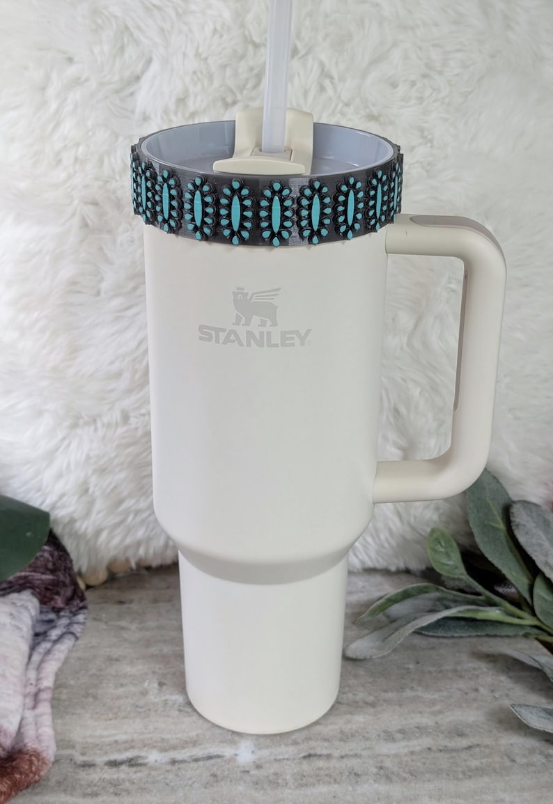 May include: A white Stanley tumbler with a handle and a decorative band featuring turquoise-colored stones. The tumbler has a straw and the Stanley logo. The overall design is clean and modern, suitable for beverages.