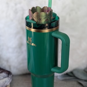 May include: A green stainless steel travel mug with a gold rim and a black handle. The mug has a logo that says "Stanley" on the side. The mug has a green and iridescent straw lid with a green straw.