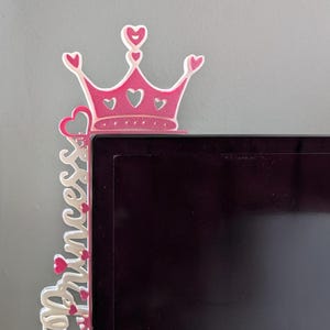 May include: A pink and white princess crown-shaped decoration. The crown has heart-shaped cutouts and is attached to a vertical piece with the word "Princess" in cursive. The decoration is placed on the corner of a black surface.