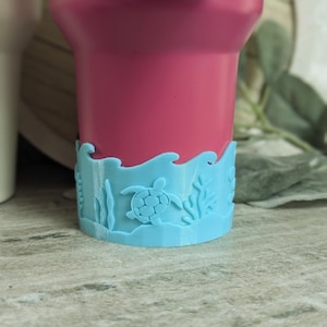 May include: A magenta tumbler with a light blue decorative base. The base features a sea turtle, seaweed, and wave design. The tumbler is shown on a light gray surface.