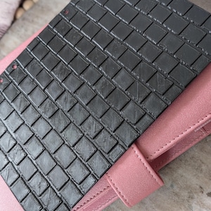 May include: A black, textured, rectangular sheet with a woven pattern, resting on a pink leather planner. The sheet has several small holes along one edge. The planner has a matching pink strap.