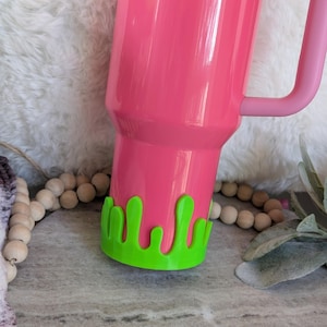 May include: A bright pink Stanley tumbler with a matching handle. The bottom of the tumbler has a green, drippy, slime-like design. The tumbler is on a gray surface with wooden beads and greenery in the background.