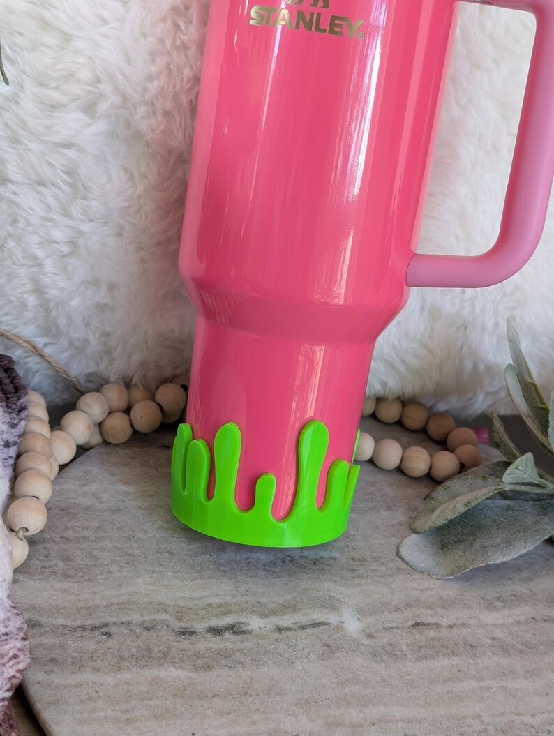 May include: A bright pink Stanley tumbler with a matching handle. The bottom of the tumbler has a green, drippy, slime-like design. The tumbler is on a gray surface with wooden beads and greenery in the background.