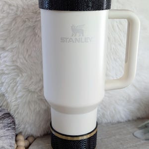 May include: Cream-colored Stanley travel mug with a black textured band and gold trim at the top and bottom. The mug has a handle and a straw. The Stanley logo is visible on the front.