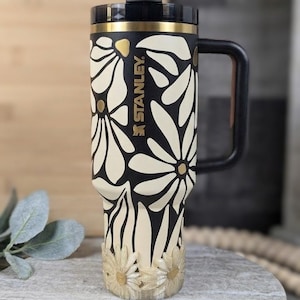 May include: A black and cream-colored Stanley travel mug with a floral design. The mug features a black handle and lid, with the Stanley logo in gold. The bottom of the mug has a decorative daisy design.