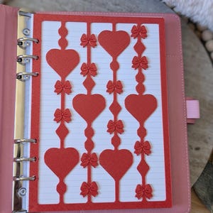 May include: A pink binder with a red decorative paper insert featuring heart and bow cutouts. The insert is placed over lined paper. The binder is open, revealing the metal rings and a pink elastic band.